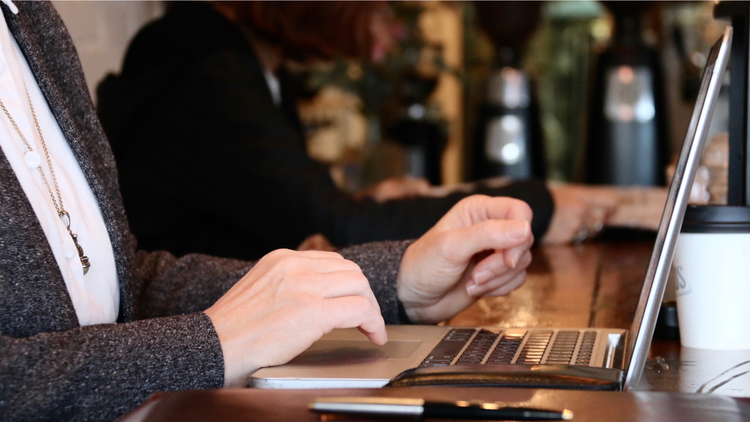 Woman in grey jacket typing on a laptop in a cafe